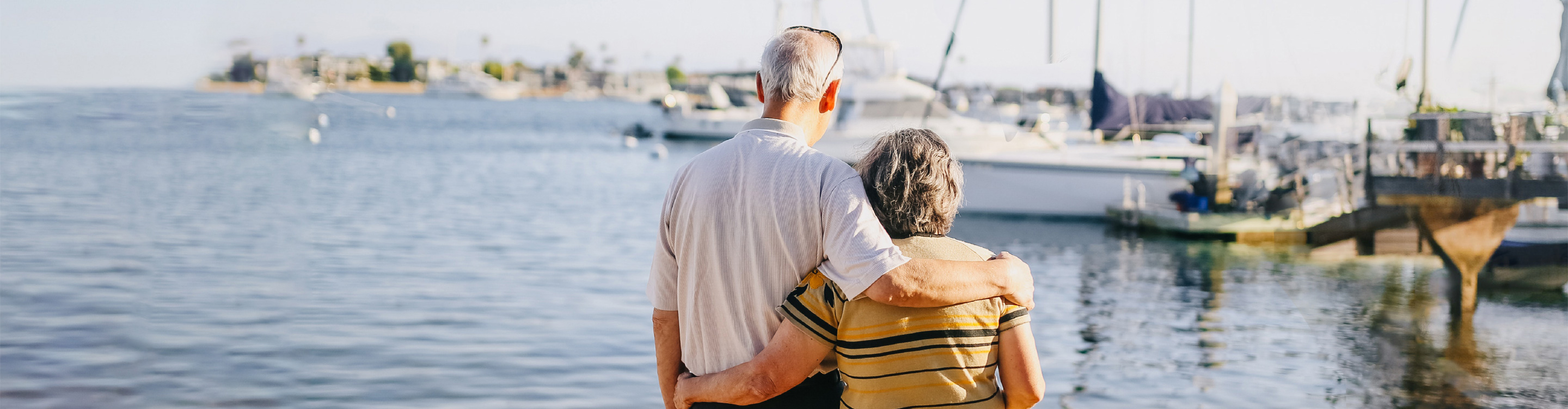Elderly man and woman with their arms around each other, looking out onto a marina. 