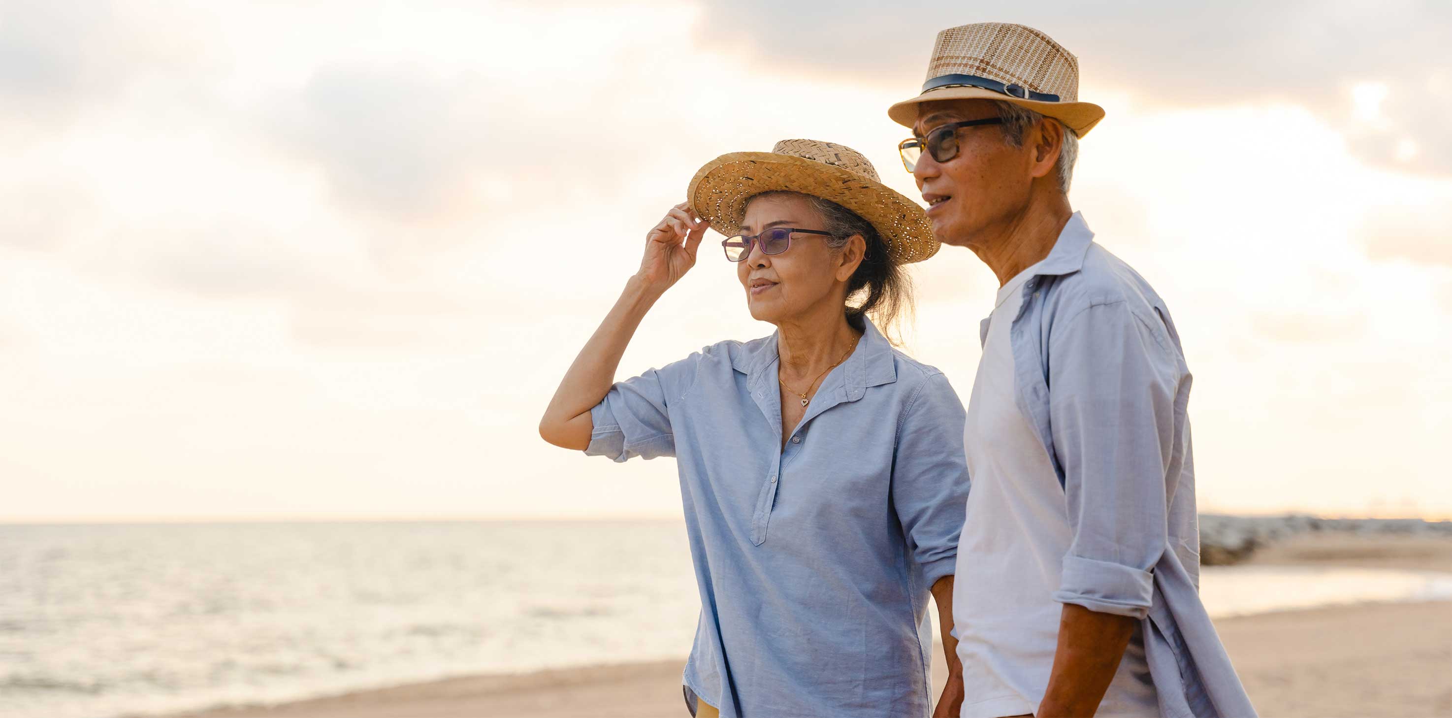 An elderly Asian couple stand on a beach looking out into the distance together. 
