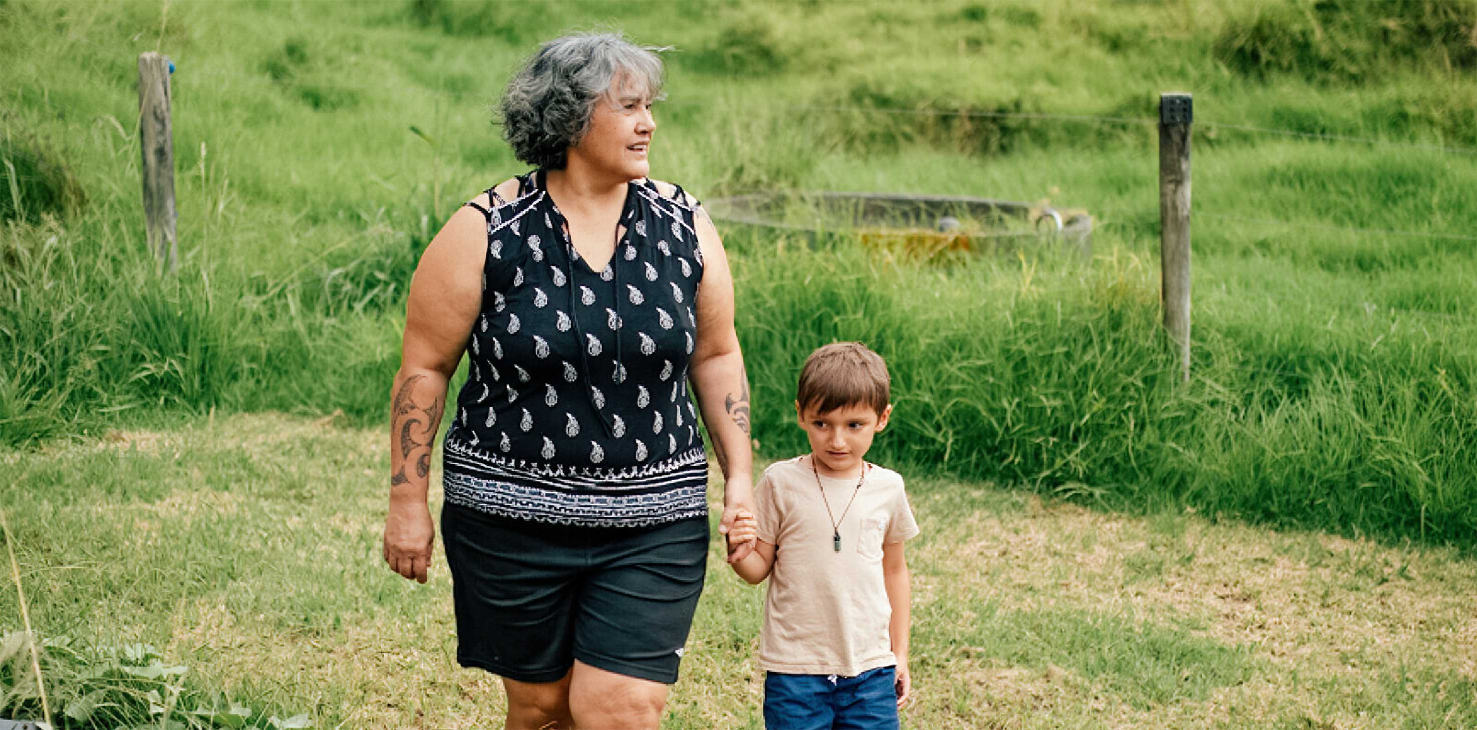 A grey-haired middle aged woman of Māori descent, walks hand in hand with her grandchild in a green paddock. 
