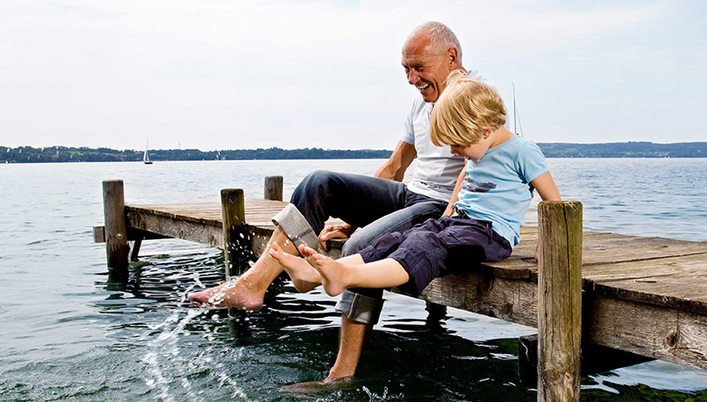 A senior man sits with his grandson on a pier, splashing the sea water with their feet.