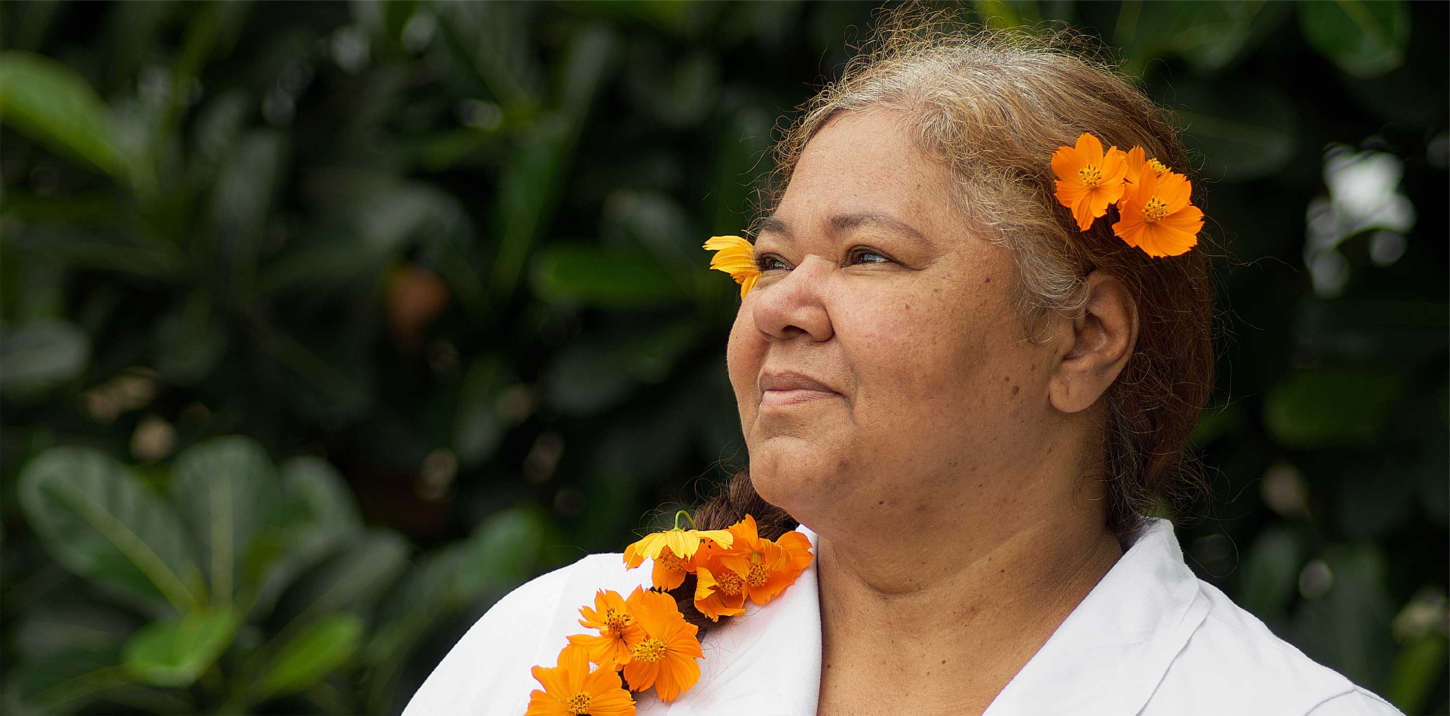 A headshot of a middle-aged Pasifika woman with orange flowers decorating her hair. She is smiling softly and gazing off into the distance.