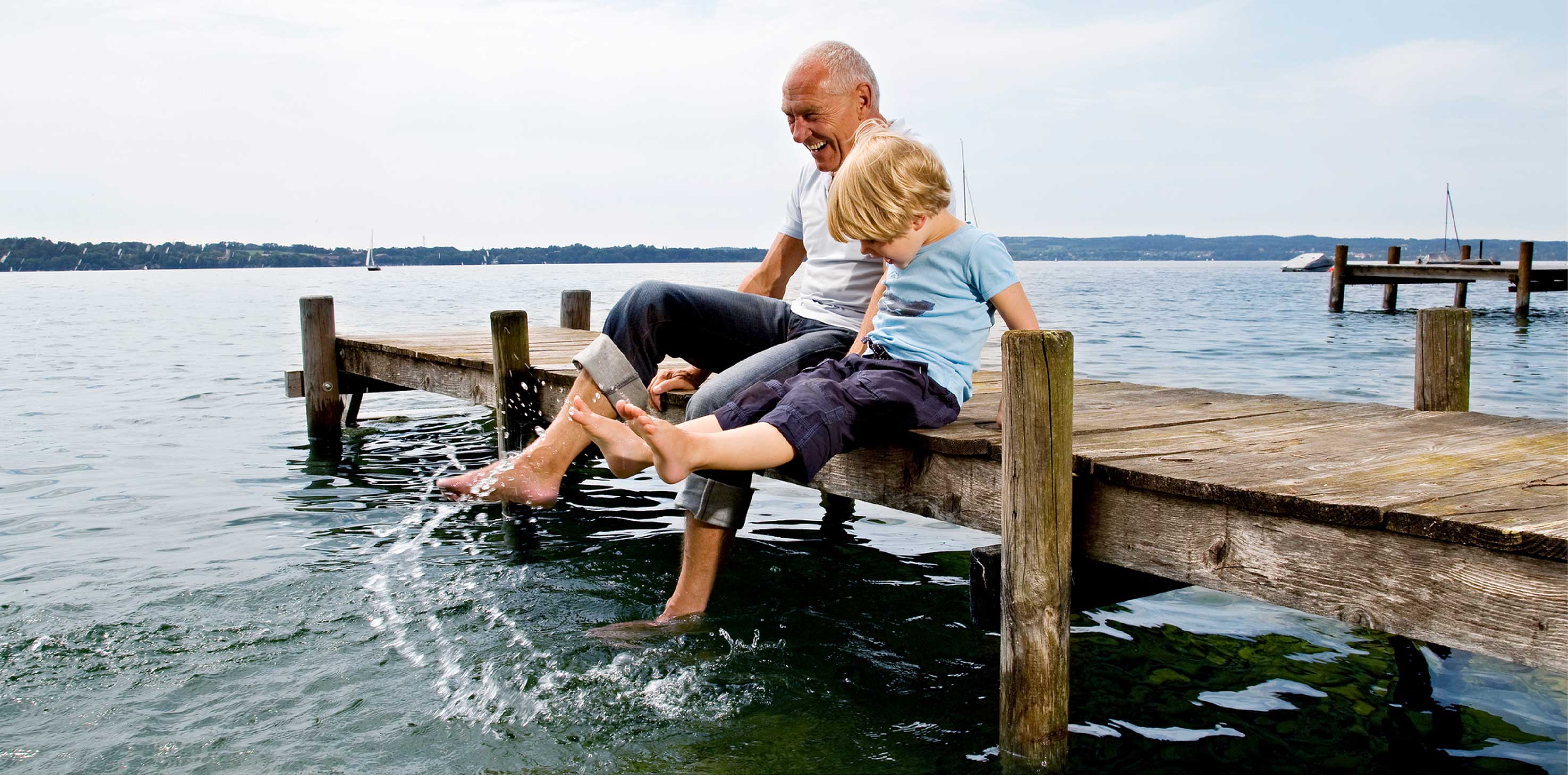 An elderly, grey-haired Caucasian man sits with his grandson on a pier, splashing the sea water with their feet.