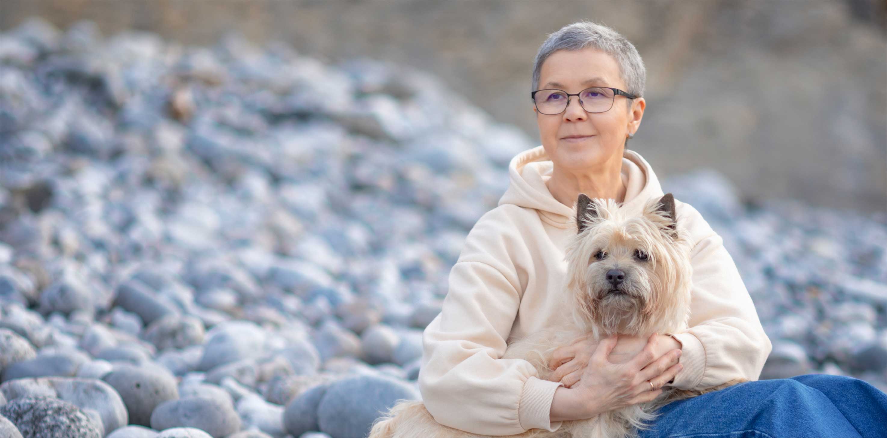 A middle aged Asian woman with grey hair sits on a pebbly beach shore with her dog in her lap. She gazes off into the distance.