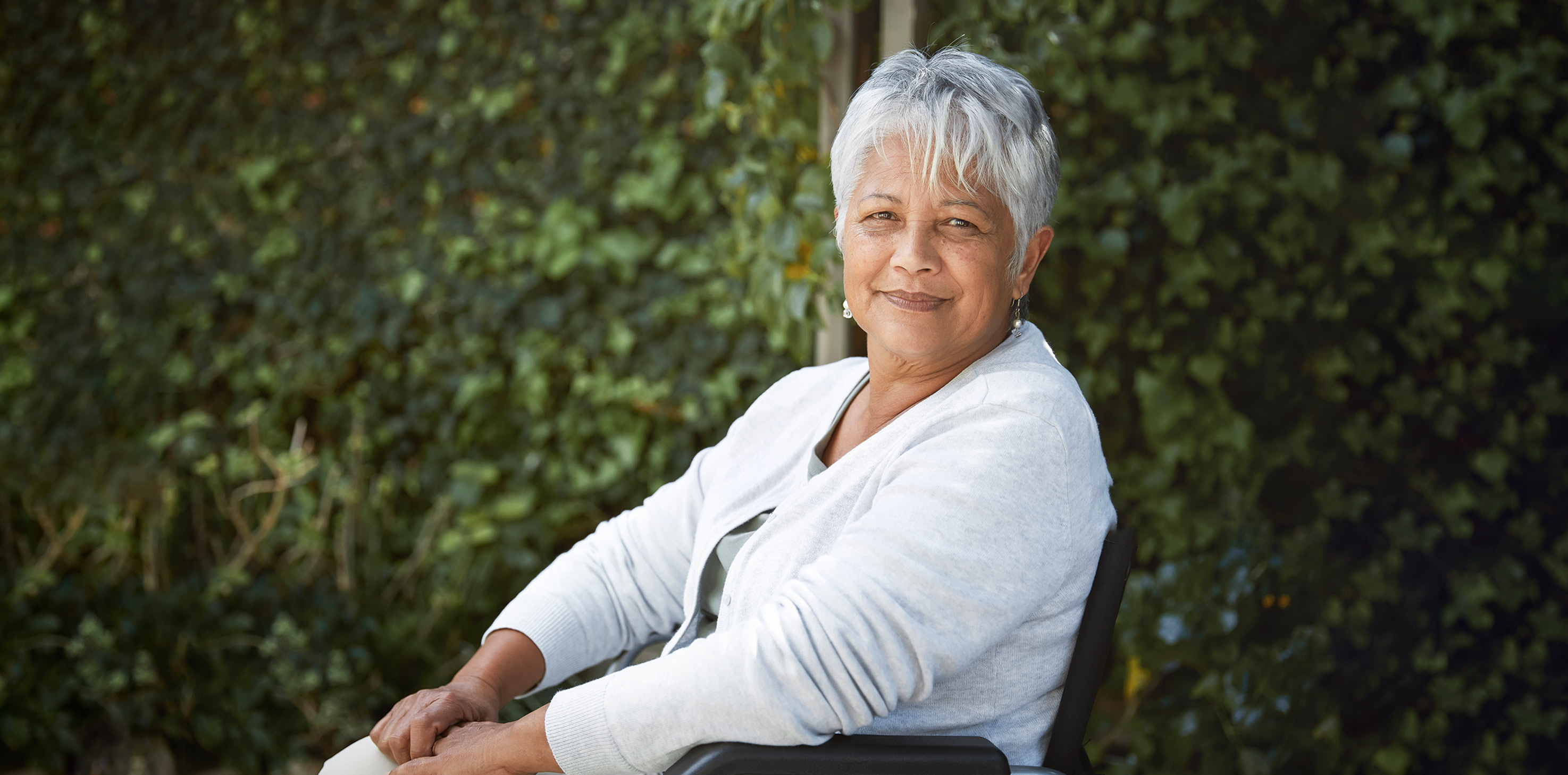 A headshot of a grey-haired woman of South Asian descent, sitting in a wheelchair and smiling softly at the camera.