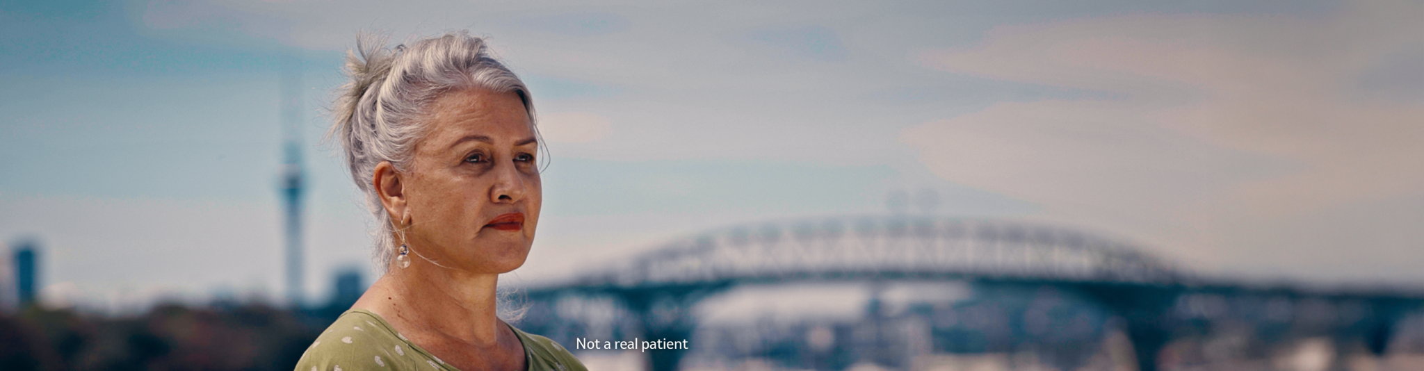 Photo of an older woman with grey hair standing outdoors in front of a blurred Auckland background. 